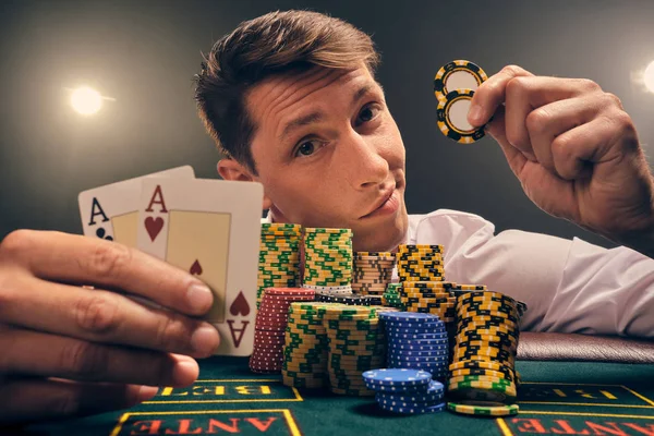 Snygg känslomässig man spelar poker sitter vid bordet i kasino mot en vit spotlight. — Stockfoto