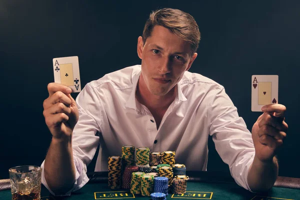 Bello uomo emotivo sta giocando a poker seduto al tavolo del casinò contro un riflettore bianco . — Foto Stock
