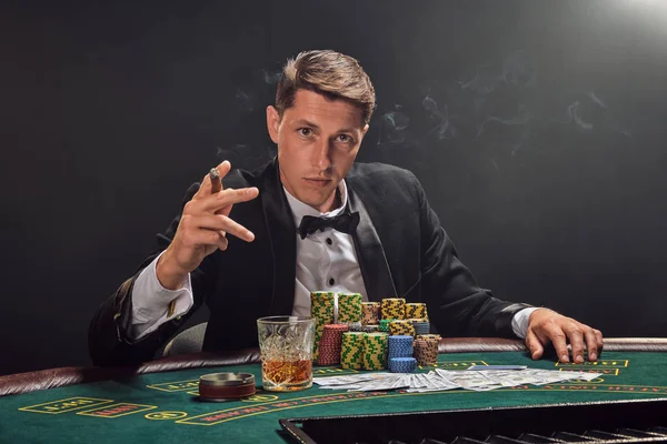 Handsome emotional man is playing poker sitting at the table in casino against a white spotlight.