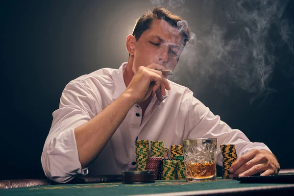 Handsome emotional man is playing poker sitting at the table in casino against a white spotlight.