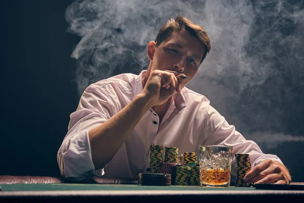 Jóképű, érzelmes férfi pókerezik az asztalnál a kaszinóban egy fehér rivaldafény ellen.. — Stock Fotó