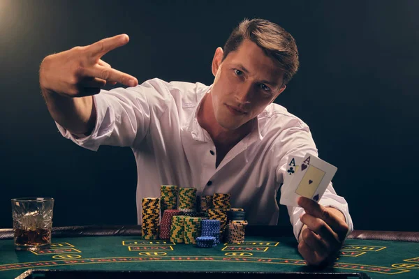 Hombre guapo emocional está jugando al póquer sentado en la mesa en el casino contra un foco blanco . —  Fotos de Stock