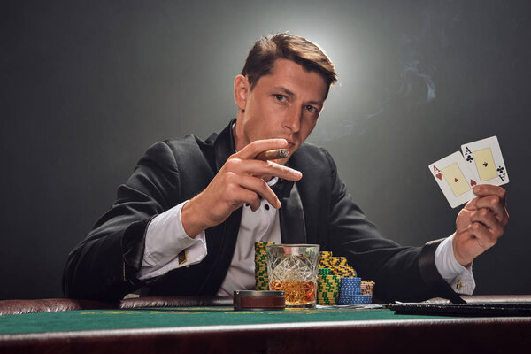 Handsome emotional man is playing poker sitting at the table in casino against a white spotlight.