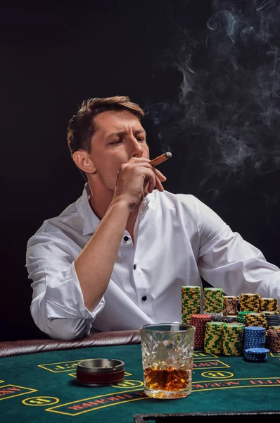 Snygg känslomässig man spelar poker sitter vid bordet i kasino mot en vit spotlight. — Stockfoto