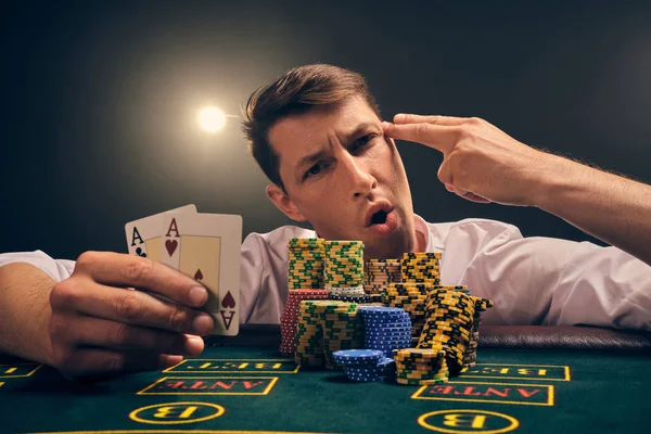Hombre guapo emocional está jugando al póquer sentado en la mesa en el casino contra un foco blanco . —  Fotos de Stock