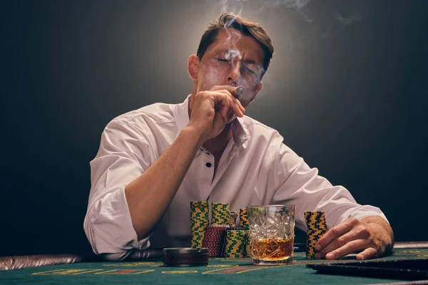 Hombre guapo emocional está jugando al póquer sentado en la mesa en el casino contra un foco blanco . — Foto de Stock