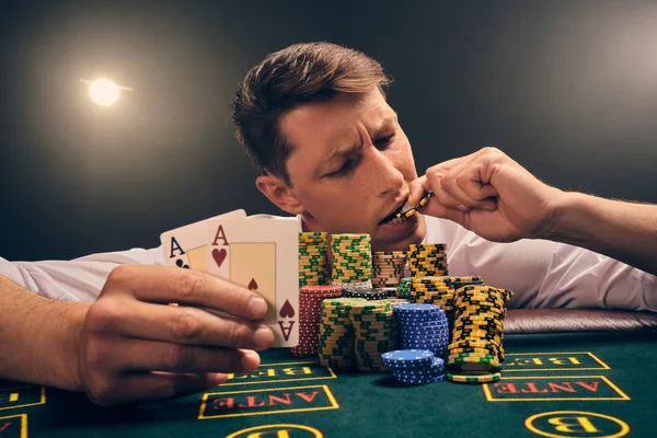 Hombre guapo emocional está jugando al póquer sentado en la mesa en el casino contra un foco blanco . — Foto de Stock