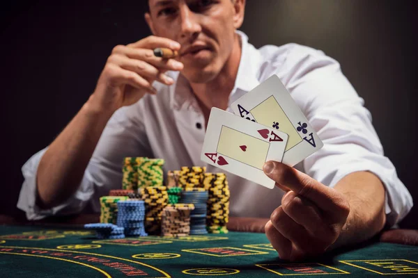 Snygg känslomässig man spelar poker sitter vid bordet i kasino mot en vit spotlight. — Stockfoto