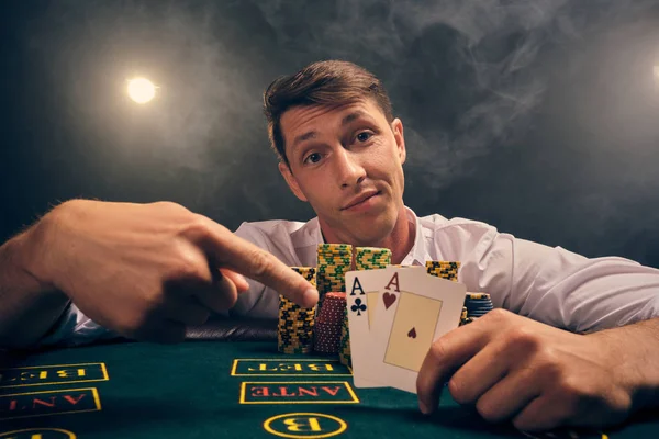 Schöner emotionaler Mann pokert am Tisch im Casino vor weißem Scheinwerferlicht. — Stockfoto