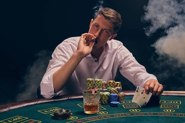 Fešák emocionální muž hraje poker sedí u stolu v kasinu proti bílému reflektoru. — Stock fotografie