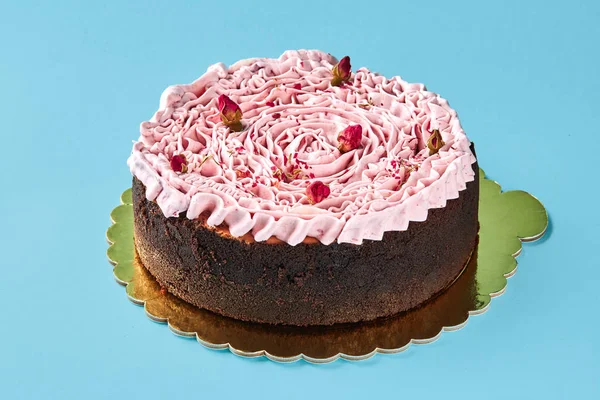 Delicious pink cream cake decorated with a small dried rosebuds, standing on a gold stand against a blue studio background. — Stock Photo, Image