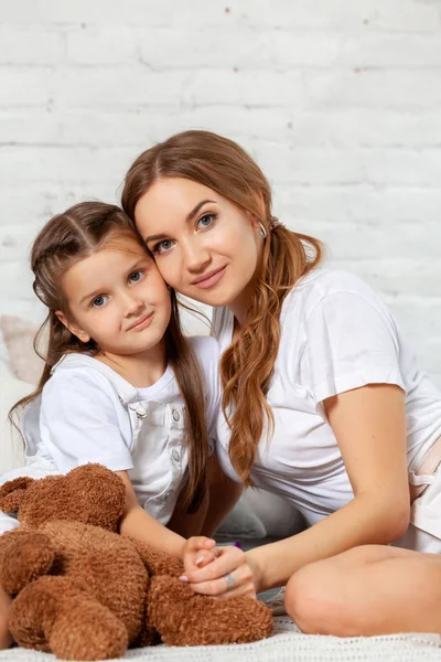 Indoor portrait of a beautiful mother with her charming little daughter posing against bedroom interior. — Stock Photo, Image