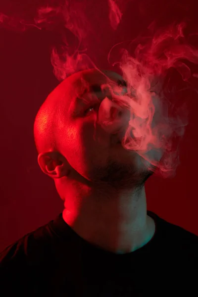 Κοντινό πλάνο στο στούντιο με ένα νεαρό φαλακρό τύπο, που ανατινάζει ένα σύννεφο καπνού σε κόκκινο φόντο. — Φωτογραφία Αρχείου