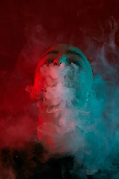 Primo piano ripresa in studio di un giovane calvo vaporizzare, soffiando una nuvola di fumo su sfondo rosso . — Foto Stock