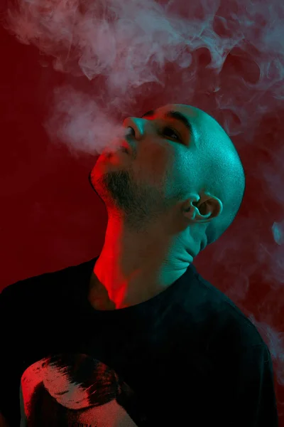 Κοντινό πλάνο στο στούντιο με ένα νεαρό φαλακρό τύπο, που ανατινάζει ένα σύννεφο καπνού σε κόκκινο φόντο. — Φωτογραφία Αρχείου