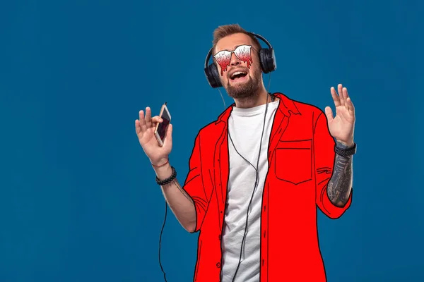 Νέος άντρας με ένα απλό πουκάμισο κινουμένων σχεδίων και γυαλιά τήξης ακούει τη μουσική και τραγουδά σε ένα μπλε φόντο. — Φωτογραφία Αρχείου