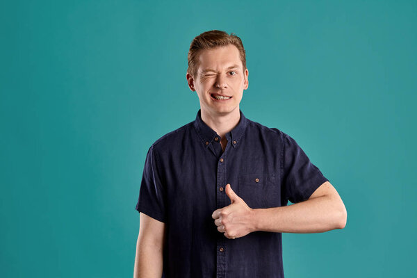 Close-up portrait of a young handsome ginger peson in a stylish navy t-shirt looking at the camera, winking and showing thumb up while posing on blue studio background. Human facial expressions. Sincere emotions concept. Copy space.