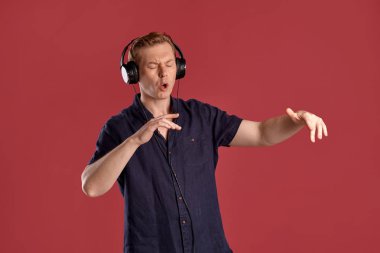 Şık bir donanma tişörtü giymiş, kulaklıkla müzik dinleyen, pembe stüdyo arka planında poz verirken şarkı söyleyen ve dans eden yakışıklı bir erkeğin yakın plan portresi. İnsan yüz ifadeleri. Samimi duygular konsepti. Boşluğu kopyala.