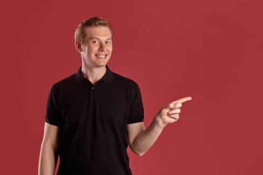 Pembe stüdyo arka planında poz verirken siyah tişörtlü, gülümseyen ve bir şeyi işaret eden görkemli bir kızıl kişinin yakın plan portresi. İnsan yüz ifadeleri. Samimi duygular konsepti. Boşluğu kopyala.