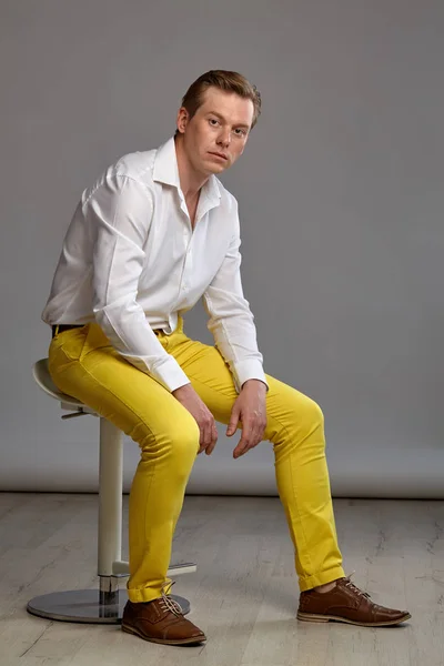 スタイリッシュな白いシャツに黄色のズボンを着た若い美しい生姜の人の完全な長さの肖像画は 灰色のスタジオの背景に対してバーチェアの上に横に座ってポーズをとっている間 カメラを見て 人間の顔の表情 誠実な感情の概念 — ストック写真