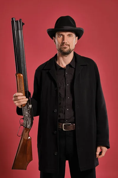 Homem de meia-idade com barba, bigode, casaco preto e chapéu, segurando uma arma enquanto posava contra um fundo vermelho. Conceito de emoções sinceras . — Fotografia de Stock