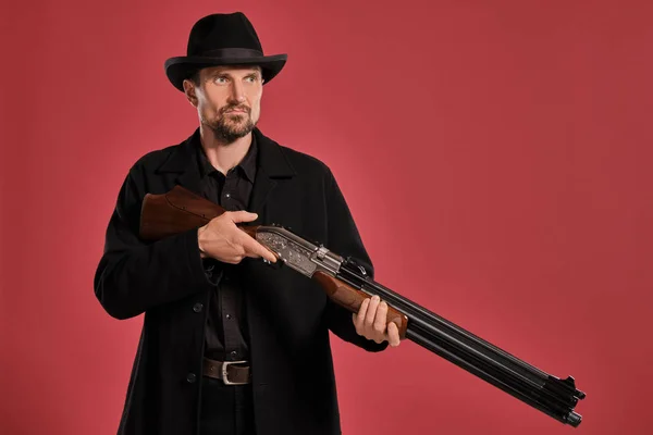 Μεσήλικα άντρας με μούσι, μουστάκι, με μαύρο μπουφάν και καπέλο, κρατώντας ένα όπλο ενώ ποζάρει σε κόκκινο φόντο. Ειλικρινή συναισθήματα έννοια. — Φωτογραφία Αρχείου