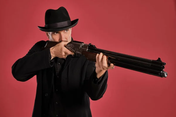 Homem de meia-idade com barba, bigode, casaco preto e chapéu, segurando uma arma enquanto posava contra um fundo vermelho. Conceito de emoções sinceras . — Fotografia de Stock