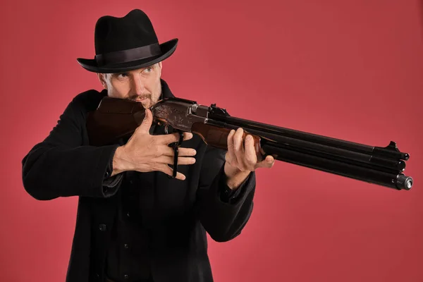 Μεσήλικα άντρας με μούσι, μουστάκι, με μαύρο μπουφάν και καπέλο, κρατώντας ένα όπλο ενώ ποζάρει σε κόκκινο φόντο. Ειλικρινή συναισθήματα έννοια. — Φωτογραφία Αρχείου