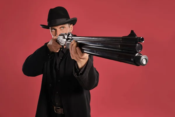 Middle-aged man with beard, mustache, in black jacket and hat, holding a gun while posing against a red background. Sincere emotions concept. — Stock Photo, Image