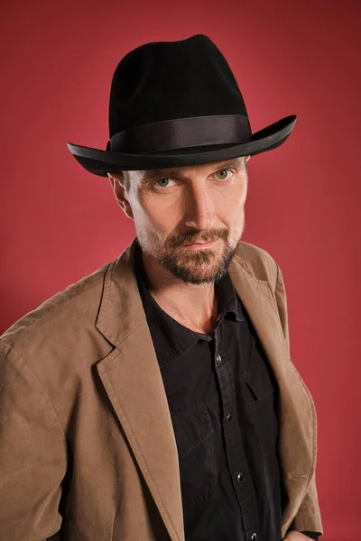 Középkorú férfi szakáll és bajusz, visel fekete kalap és barna kabát pózol ellen piros háttér. Őszinte érzelmek fogalma. — Stock Fotó