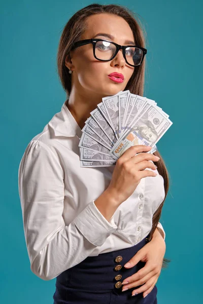 Brunetka dziewczyna w okularach, ubrana w czarną krótką spódnicę i białą bluzkę pozuje gospodarstwa fan stu rachunków dolara na niebieskim tle. — Zdjęcie stockowe