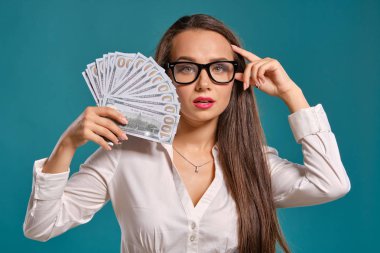 Gözlüklü esmer kız, siyah kısa etek ve beyaz bluz giyen mavi bir arka plan karşı yüz dolarlık banknotlar bir fan tutarak poz.