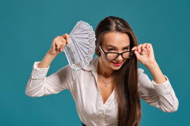 Gözlüklü esmer kız, siyah kısa etek ve beyaz bluz giyen mavi bir arka plan karşı yüz dolarlık banknotlar bir fan tutarak poz.