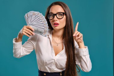 Gözlüklü esmer kız, siyah kısa etek ve beyaz bluz giyen mavi bir arka plan karşı yüz dolarlık banknotlar bir fan tutarak poz.