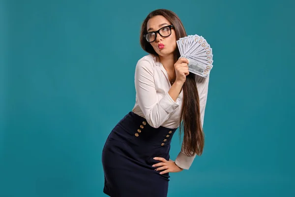 Brunette girl in glasses, wearing in a black short skirt and white blouse is posing holding a fan of hundred dollar bills against a blue background. — Stock Photo, Image