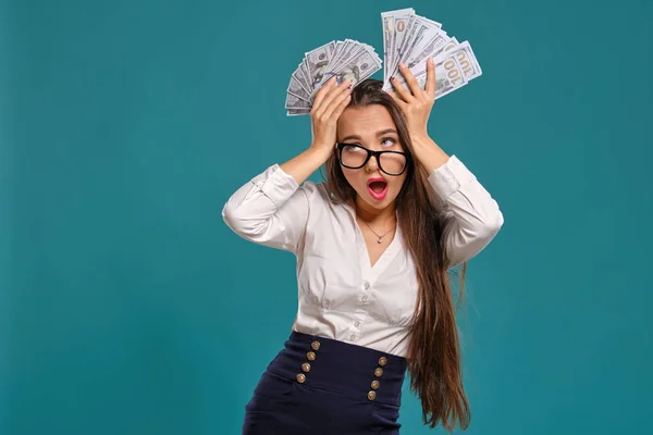 Brunette girl in glasses, wearing in a black short skirt and white blouse is posing holding a fan of hundred dollar bills against a blue background.