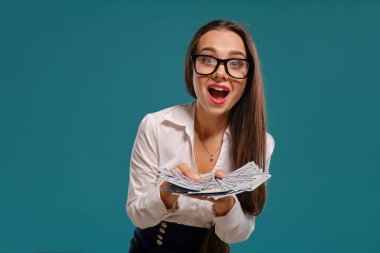 Gözlüklü esmer kız, siyah kısa etek ve beyaz bluz giyen mavi bir arka plan karşı yüz dolarlık banknotlar bir fan tutarak poz.