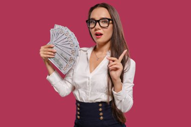 Gözlüklü esmer kız, siyah kısa etek ve beyaz bluz giyen pembe bir arka plan karşı yüz dolarlık banknotlar bir fan tutarak poz.