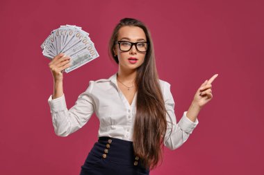Gözlüklü esmer kız, siyah kısa etek ve beyaz bluz giyen pembe bir arka plan karşı yüz dolarlık banknotlar bir fan tutarak poz.
