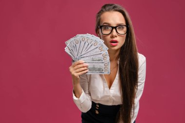 Gözlüklü esmer kız, siyah kısa etek ve beyaz bluz giyen pembe bir arka plan karşı yüz dolarlık banknotlar bir fan tutarak poz.