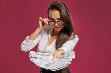 Gözlüklü esmer kız, siyah kısa etek ve beyaz bluz giyen pembe bir arka plan karşı yüz dolarlık banknotlar bir fan tutarak poz.