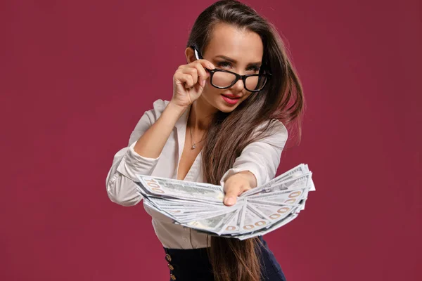 Menina morena em óculos, vestindo em uma saia curta preta e blusa branca está posando segurando um fã de notas de cem dólares contra um fundo rosa . — Fotografia de Stock