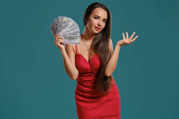 Brunetka s dlouhými vlasy, oblečené do sexy červených šatů, se na ní drží fanoušek stovek dolarových bankovek na modrém pozadí. — Stock fotografie