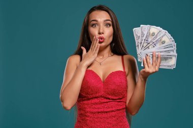 Uzun saçlı esmer kız, seksi kırmızı bir elbise giyen mavi bir arka plan karşı yüz dolarlık banknotlar bir fan tutarak poz.