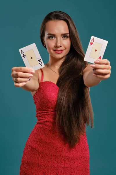 Brunette girl with a long hair, wearing a sexy red dress is posing holding two playing cards in her hands, blue background.