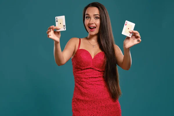 Brunette girl with a long hair, wearing a sexy red dress is posing holding two playing cards in her hands, blue background.