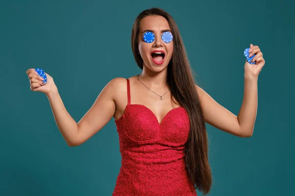 Menina morena com um cabelo longo, vestindo um vestido vermelho sexy está segurando algumas fichas de jogo em suas mãos enquanto posando contra um fundo azul . — Fotografia de Stock