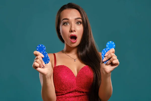 Brunette fille aux cheveux longs, portant une robe rouge sexy tient quelques jetons de jeu dans ses mains tout en posant sur un fond bleu . — Photo