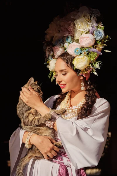 Brunett tjej i en vit ukrainsk autentisk nationell kostym och en krans av blommor poserar för kameran. Närbild. — Stockfoto