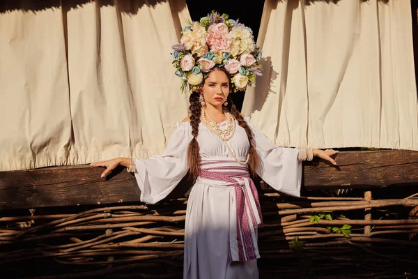 Barna lány egy fehér ukrán hiteles nemzeti ruha és a koszorú virágok pózol ellen terasz. — Stock Fotó